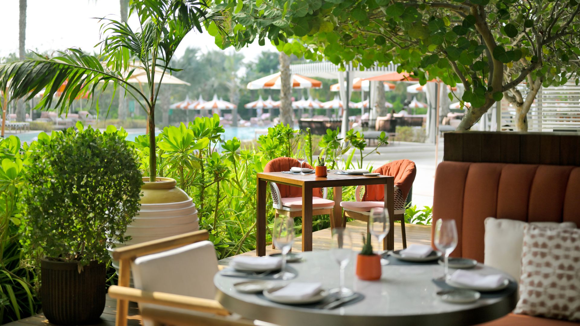 A stylish outdoor restaurant seating area surrounded by lush greenery, overlooking a poolside with sun loungers and umbrellas. The dining setup includes elegantly set tables with napkins, wine glasses, and small potted plants, creating a tranquil and inviting ambiance.